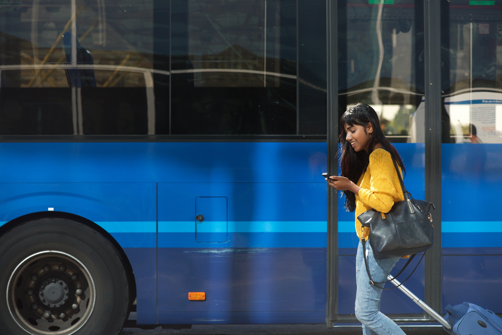 smiling young Indian woman walking with suitcase and cellphone by bus station