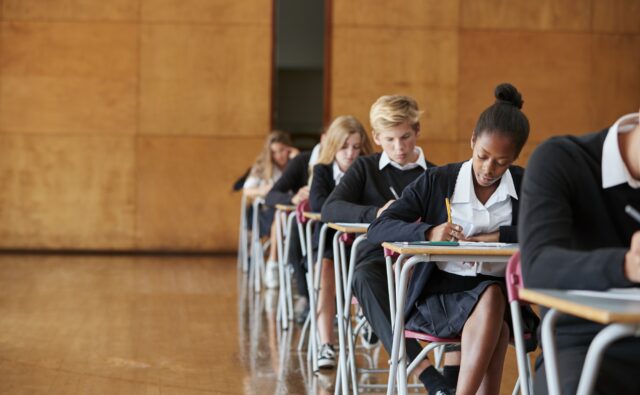 Teenage Students In Uniform Sitting Examination In School Hall