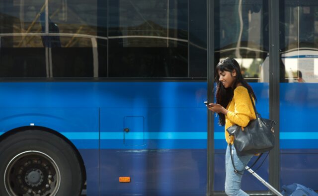 smiling young Indian woman walking with suitcase and cellphone by bus station